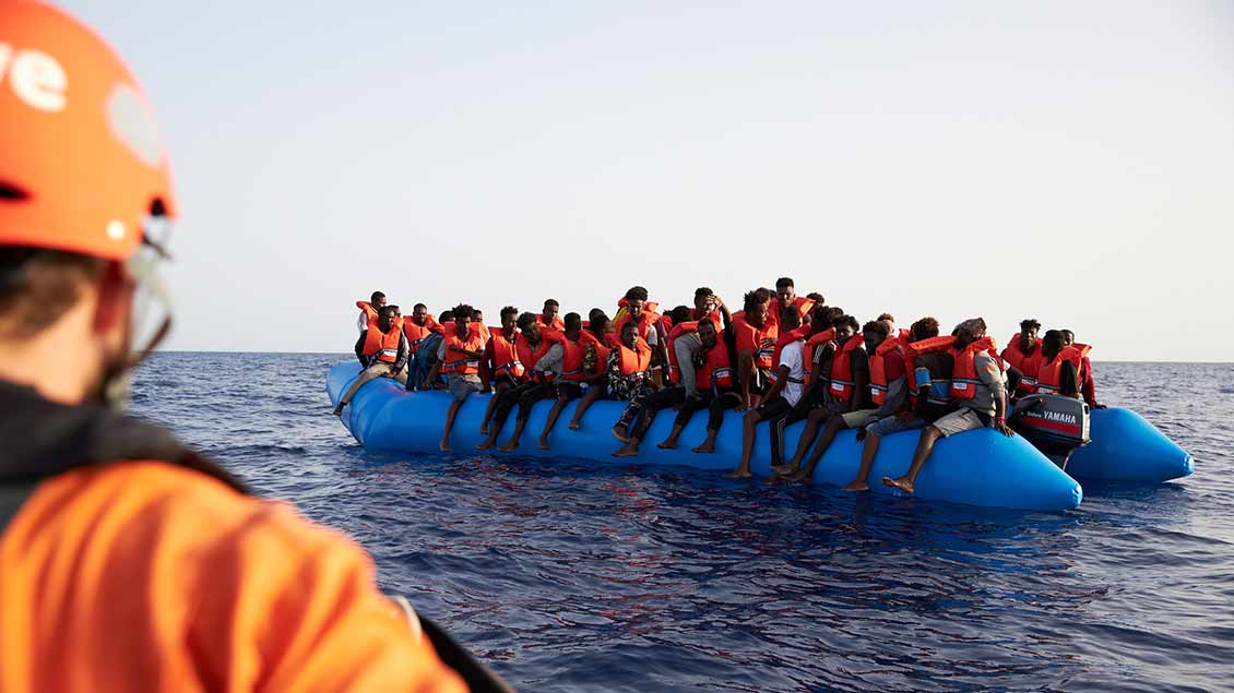 Migranten auf einem Schlauchboot, im Vordergrund ein Mitarbeiter der Organisation Sea-Eye.